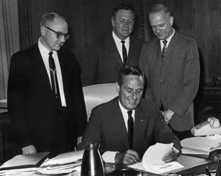 male signing document at desk with three other males standing around him